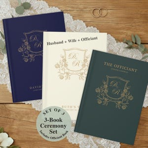May include: A three-book ceremony set with navy, cream, and teal covers. Each book features a gold crest design with floral accents and personalized text. The set includes books for the husband, wife, and officiant. The set is displayed on a wooden surface.