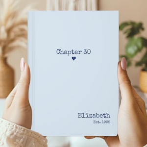 Puede incluir: Un libro azul claro con el texto "Chapter 30" encima de un pequeño corazón azul. Debajo, el nombre "Elizabeth" está impreso, seguido de "Est. 1995". El libro está sostenido por una persona.