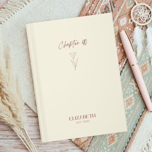 May include: A cream-coloured journal with the text "Chapter 40" and the name "ELIZABETH EST. 1985" in burgundy script. A matching burgundy pen rests beside the journal. The journal features a floral illustration.