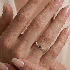May include: A gold band ring featuring a series of small, dark blue four-petalled flower motifs alternating with tiny clear stones. The ring is thin and delicate, encircling the finger with a repeating pattern of these floral and stone elements.
