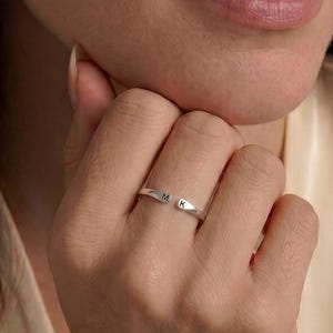 Puede incluir: Un anillo abierto de tono plateado con dos extremos planos y cuadrados. Cada extremo está grabado con una letra mayúscula, las letras M y K, posicionadas de manera que quedan orientadas hacia afuera en el dedo.