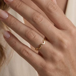Puede incluir: Un anillo fino de oro que presenta un patrón continuo de ondas o zigzag a lo largo de la banda. El anillo se lleva en el dedo anular y muestra un acabado metálico pulido que refleja la luz sobre su estructura de diseño ondulado.