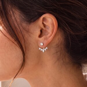 May include: A silver-toned ear jacket earring featuring a small round stud at the front and a curved, fan-shaped attachment behind the earlobe. The attachment is adorned with five clear, faceted stones in varying shapes, including round and pear cuts.