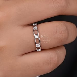 Op de afbeelding: Een zilveren ring met een reeks kleine, vierkant geslepen diamanten in een geschakeld ontwerp. De ring zit om een vinger met donkerrode nagellak. De achtergrond is donker.