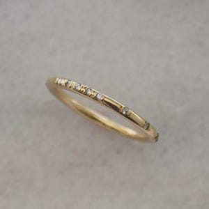 Könnte beinhalten: Ein dünner Goldring mit einer Reihe kleiner, gleichmäßig verteilter, klarer Steine, die entlang der oberen Hälfte des kreisförmigen Metallbandes gefasst sind. Der Ring hat eine polierte Oberfläche und ein schlichtes, schmales Profil.