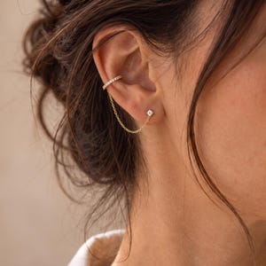 May include: A gold-toned ear cuff featuring a row of small clear stones connected by a delicate fine chain to a single stud earring with a clear stone. The jewelry is worn on the upper cartilage and the earlobe, creating a linked chain effect.