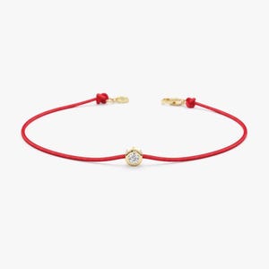 May include: A red string bracelet with a gold clasp and a small, round, gold charm with a single, clear gemstone in the center. The bracelet is simple and elegant, suitable for everyday wear.