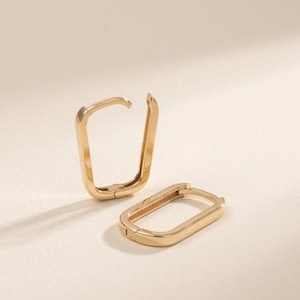 Könnte beinhalten: Zwei goldfarbene, rechteckige Creolen. Ein Ohrring ist offen, der andere geschlossen. Die Ohrringe befinden sich auf einer hellen Oberfläche. Die Ohrringe haben ein schlichtes, modernes Design.