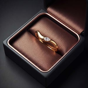May include: A gold ring with a smooth, curved band featuring a single marquise-cut clear gemstone set in the centre. The ring is displayed inside a black jewellery box with a brown satin interior lining that matches the ring's metallic tone.