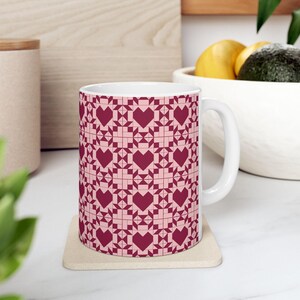 May include: White ceramic mug with a repeating pattern of burgundy hearts and geometric shapes on a light pink background. The mug has a white handle and sits on a beige coaster. The design resembles a quilt pattern.