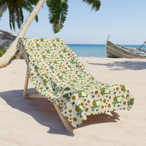 May include: A beach towel featuring a repeating pattern of green frogs, brown circular shapes, and small clusters of dark blue dots on a light cream background, draped over a wooden lounge chair on a sandy beach near the ocean.