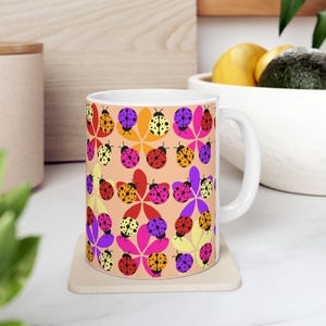 Puede incluir: Taza de cerámica blanca con un patrón repetido de mariquitas y formas de flores coloridas. Las mariquitas son de varios colores, incluyendo rojo, morado, amarillo y naranja, sobre un fondo melocotón. La taza está sobre un posavasos cuadrado.