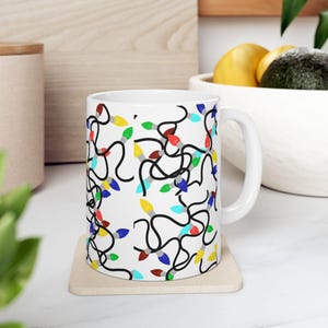 May include: White ceramic mug featuring a repeating pattern of colorful Christmas lights with black cords. The lights are in various colors, including red, yellow, green, and blue. The mug has a white handle and is sitting on a beige coaster.