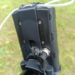 May include: A black electronic device with a textured surface, secured by a black strap and mounted on a black pole. A white cord is attached to the device. The device has a connector and a cable.
