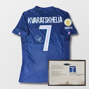 Pode incluir: Uma camisola de futebol azul marinho com o nome "KVARATSKHELIA" e o número "7" em branco. A camisola tem uma assinatura e um patch "LIGUE 1". Um certificado emoldurado é exibido ao lado da camisola.
