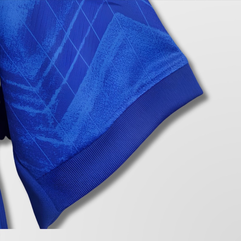 Puede incluir: Primer plano de una manga de camiseta deportiva azul real. La manga presenta un patr&oacute;n texturizado y un pu&ntilde;o acanalado. La tela tiene un brillo sutil y el dise&ntilde;o incluye un patr&oacute;n geom&eacute;trico.