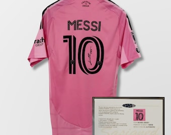 Puede incluir: Camiseta de fútbol rosa con el nombre "MESSI" y el número "10" en negro. La camiseta tiene una firma y las palabras "FREEDOM DREAM" en la espalda. Se muestra un certificado enmarcado al lado.