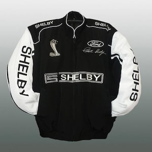 Puede incluir: Una chaqueta negra y blanca con cremallera que presenta la palabra SHELBY impresa en las mangas y el pecho. El frente muestra un emblema de cobra, un logotipo de Ford y la firma de Carroll Shelby. Incluye paneles blancos en las mangas.