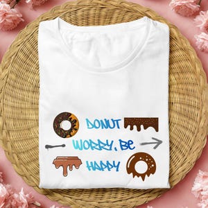 May include: White t-shirt with a donut-themed graphic. The design includes donut illustrations and the phrase "Donut Worry, Be Happy" in blue and brown colors. The shirt is folded on a woven basket.
