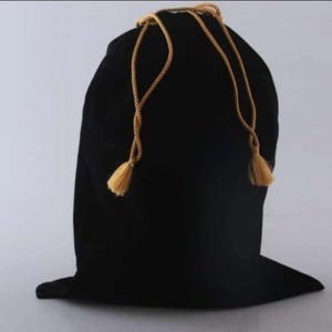 Peut inclure: Un sac en velours noir avec cordon de serrage et glands dorés. Le sac est fermé en haut par le cordon de serrage. Le sac est debout sur une surface blanche.
