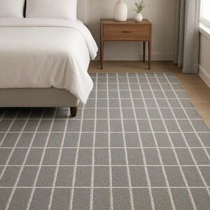 Puede incluir: Una alfombra rectangular gris con un patrón de cuadrícula en un dormitorio. La alfombra tiene un aspecto suave y texturizado con un fondo gris claro y líneas de cuadrícula blancas. El dormitorio incluye una cama, una mesita de noche y una lámpara.