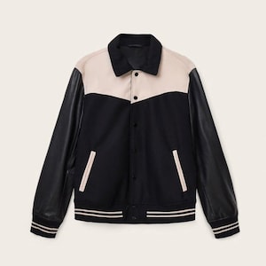 Può includere: Una giacca college nera e beige con maniche in pelle nera. La giacca ha un colletto e un pannello superiore del petto beige, con un petto e un corpo inferiori neri. Presenta due tasche anteriori e polsini e orlo a righe.