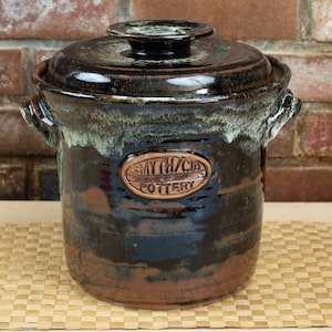 Op de afbeelding: Een bruine en zwarte keramische pot met een deksel. De pot heeft een witte glazuur en een koperkleurig etiket met de tekst "Myth/Cid Pottery".