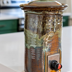 Puede incluir: Un dispensador de vino de cerámica con un esmalte marrón, verde y dorado. El dispensador tiene un pico con un tapón negro y rojo.