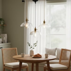 Puede incluir: Tres lámparas colgantes con pantallas de vidrio transparente y festoneado y herrajes de color latón cuelgan sobre una mesa redonda de madera. La habitación tiene una paleta de colores neutros con una ventana y dos sillas con respaldos tejidos.
