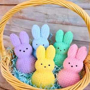 May include: A woven basket filled with five colorful bunny-shaped candies. The bunnies are in shades of purple, blue, green, yellow, and pink, each with black dot eyes. The basket contains blue paper grass.