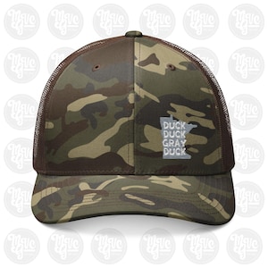 May include: A camouflage print trucker hat with brown mesh back panels and a brown button on top. The front panel features an embroidered white outline of the state of Minnesota with the text DUCK DUCK GRAY DUCK stacked inside the shape.