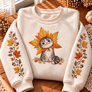 Può includere: Una felpa girocollo color crema con un ricamo raffigurante un gattino grigio tigrato incorniciato da una grande foglia d'acero arancione. Le maniche sono decorate con tralci ricamati di foglie autunnali, ghiande e bacche rosse.