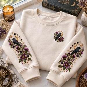 May include: A cream-colored crewneck sweatshirt featuring detailed embroidery on both sleeves and the chest. The design includes black crows, purple thistle flowers, blackberries, golden crescent moons, and celestial star motifs.