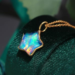 Puede incluir: Un colgante en forma de estrella con una piedra de ópalo iridiscente vibrante con destellos azules, verdes y naranjas, engastado en un fino marco de metal dorado. El colgante está unido a una delicada cadena de eslabones dorados.