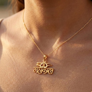Può includere: Una collana con pendente dorato con una catena sottile. Il pendente presenta un design stilizzato e la parola "महाकाल" in un carattere script. La collana è indossata intorno al collo.