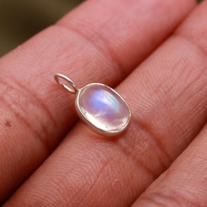 Può includere: Un piccolo ciondolo ovale con una pietra di luna iridescente lucidata incastonata in una cornice d'argento. La pietra di luna mostra una morbida, eterea lucentezza con sfumature di blu e bianco. Il ciondolo ha un piccolo anello per una catena. Circa 1,5 cm di lunghezza.