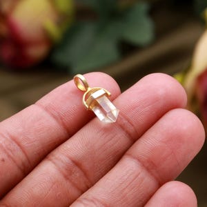 May include: A small, clear quartz crystal pendant with a gold-colored metal setting. The crystal is faceted and has a hexagonal shape. The pendant is held between fingers, with a blurred background of green and pink.