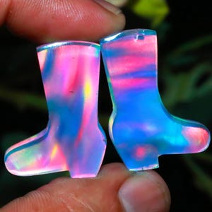 Puede incluir: Dos colgantes de bota iridiscentes en miniatura. Las botas, sujetas entre los dedos, muestran una vibrante gama de colores, incluyendo rosa, azul y amarillo. Los colgantes probablemente están hechos de un material translúcido, que atrapa la luz y crea un efecto brillante.
