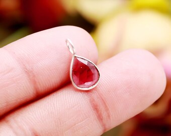 May include: A teardrop-shaped pendant featuring a faceted, deep red gemstone set in a silver bezel. The pendant is suspended from a small silver loop, suitable for a necklace or bracelet. The gemstone has a polished, reflective surface.