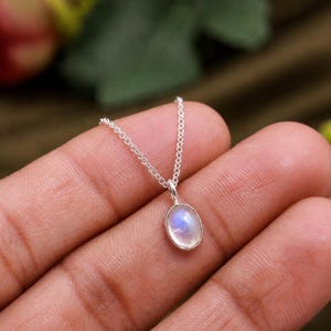 May include: A delicate silver chain necklace featuring a small, oval-shaped moonstone pendant. The stone displays a soft, iridescent blue glow within a smooth, polished silver bezel setting, suspended from a fine, lightweight silver link chain.