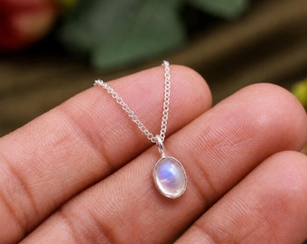 May include: A delicate silver chain necklace featuring a small, oval-shaped moonstone pendant. The stone displays a soft, iridescent blue glow within a smooth, polished silver bezel setting, suspended from a fine, lightweight silver link chain.