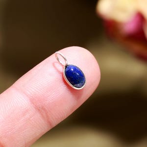 May include: A small, oval pendant featuring a deep blue gemstone set in a silver-coloured metal. The gemstone is smooth and polished, and the pendant has a small loop. The blurred background draws attention to the pendant.