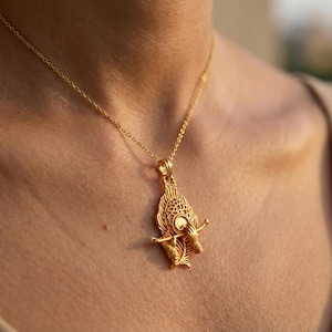 Puede incluir: Un collar con colgante dorado con un diseño ornamentado y detallado. El colgante presenta un elemento central circular con patrones intrincados y una figura que sostiene un arco y una flecha. La delicada cadena complementa el diseño del colgante.