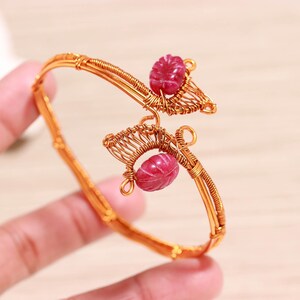 May include: A gold-coloured wire bracelet with two carved, oval-shaped, red gemstones. The bracelet features detailed wirework, creating a unique accessory. The gemstones are a vibrant red colour. The bracelet is a piece of jewellery.