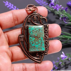Puede incluir: Colgante de cobre con una piedra turquesa rectangular en el centro. El colgante tiene un diseño ornamental en forma de escudo con detalles intrincados y una fianza de color cobre para colgar. La piedra tiene un aspecto natural moteado.