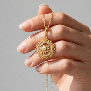 以下が含まれることがあります： 太陽のデザインが施されたゴールドカラーのペンダントネックレス。ペンダントの中央には太陽の顔が詳細に描かれ、小さな透明な石のリングとテクスチャ加工された外側の縁で囲まれています。ネックレスは、柔らかい灰色の背景に対して手で持たれています。