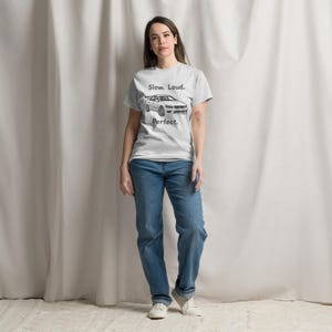 Puede incluir: Camiseta gris claro con el texto "Slow. Loud. Perfect." y un gráfico de un coche. La persona lleva vaqueros azules y zapatillas blancas.