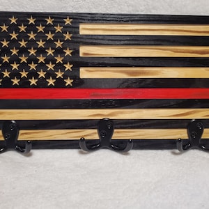 May include: Wooden wall-mounted coat rack featuring a distressed American flag design with a red stripe. The rack has three black metal hooks for hanging items. The stars are a natural wood color, and the stripes are black and natural wood.