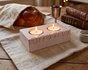 May include: A rectangular light pink stone candle holder with two circular cutouts for tea lights. The Hebrew text "Shabbat Shalom" is carved into the front face. Two lit white tea light candles are placed inside the stone candle holder.