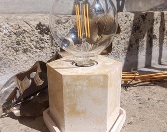 May include: A table lamp featuring a hexagonal concrete base with a mottled cream and yellow finish, resting on a matching hexagonal tray. A large, clear, spherical glass light bulb with visible vertical yellow filaments sits on top.